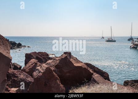 Rote Felsen an einem Inselstrand und Blick auf Segelboote und den Horizont weit voraus Stockfoto