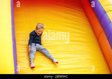 Lachend, viel Spaß, kleiner Junge, der auf einer aufblasbaren mehrfarbigen Rutsche gleitet. Hellgelber Hintergrund, freier Platz für Text. Stockfoto
