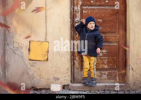 Ein kleiner Junge posiert an einem Herbsttag vor der uralten Tür und der rissigen Wand in der Altstadt. Der herbstliche Wind bläst die bunten Blätter der Bäume auf, Stockfoto