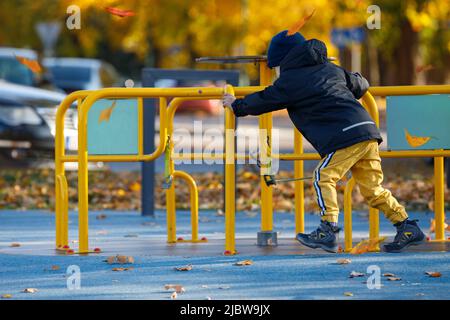 Der kleine Junge spielt auf dem Spielplatz im Park. Kind dreht ein gelbes Karussell. Herbstsaison mit leuchtend gelben Bäumen im Hintergrund. Vertikaler ph-Wert Stockfoto