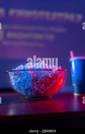 Popcorn in einer Glasschüssel und ein Limonadengetränk in einem Glas. Nahaufnahme. Sorgenfreies Betrachten von Fernsehprogrammen, Lieblingsfilmen. Urlaubssymbole. Neonbeleuchtung Stockfoto