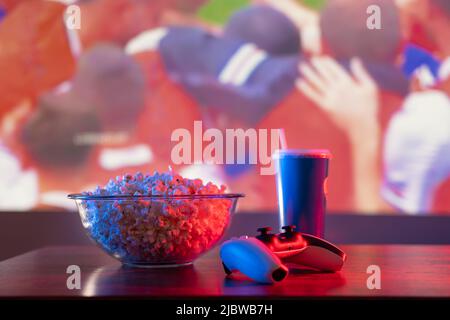 Popcorn in einer Schüssel, ein Glas Sodawasser mit einem Stroh und ein Gamepad auf einem romantischen Hintergrund. Nahaufnahme. Sehen Sie Ihre Lieblingsvideos auf dem Fernseher, Video Gam Stockfoto