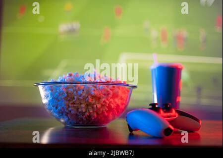 Ein Glas Limonade mit einem Strohhalm, eine Glasschüssel Popcorn, ein Gamepad auf einem sportlichen Hintergrund. Videospiele auf dem großen Fernsehbildschirm, Wettbewerbe mit fr Stockfoto