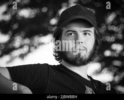 Junger erwachsener Mann mit längeren dunklen Haaren, dunklen Augen, dunklem Bart und Schnurrbart mit einer Baseballmütze, der im Sommer im Park sitzt und darüber nachdenkt Stockfoto