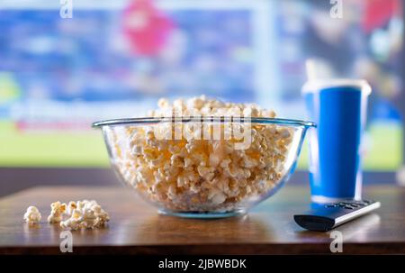 Nahaufnahme. Transparente Glasschüssel mit köstlichem Popcorn. Im Hintergrund befindet sich ein blaues Glas und eine Fernbedienung auf dem Tisch. TV-Hintergrund. Ansehen Stockfoto