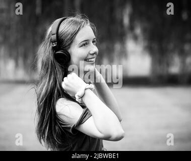 Junge langhaarige Teenager-Mädchen mit Ohrhörern lächeln auf einem Rasen aus Gras mit einer Weide im Hintergrund im Sommer in schwarz und weiß Stockfoto