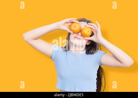 Junge kaukasische Mädchen isoliert auf orangefarbenem Hintergrund bedeckt ihre Augen mit zwei orangefarbenen Früchten. Leckere Früchte reich an Vitamin c Stockfoto