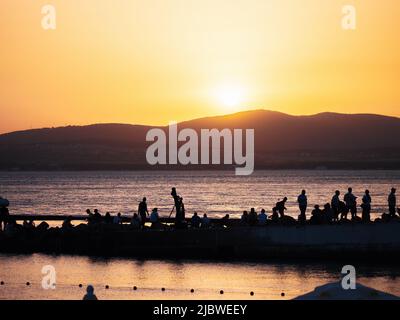 Silhouetten vieler Menschen am Pier in der Meeresbucht vor dem Hintergrund des Sonnenuntergangs und der Berge. Summertime and Relax-Konzept mit Kopierbereich. Stockfoto