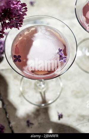 Champagner-Rosé-Glas mit Fliederblumen und Schatten auf grauem Hintergrund, Nahaufnahme, Platz für Text. Sommerdrink für Party, Weinladen oder Verkostungsconcep Stockfoto