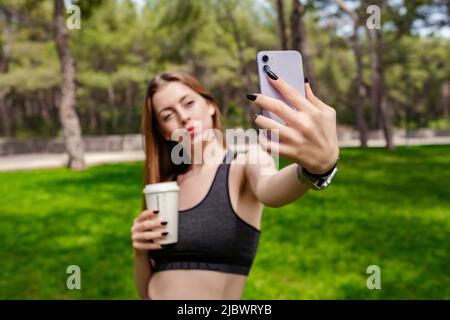 Glückliche Brünette Frau trägt schwarzen Sport-BH auf Stadtpark stehen, im Freien Selfie und hält Takeaway Kaffeebecher. Selbstporträt für soziale Medien Stockfoto