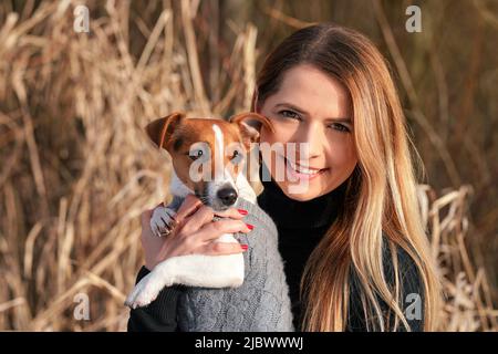 Junge Frau, die ihren Jack Russell Terrier Hund in warmer Winterkleidung an den Händen hält Stockfoto