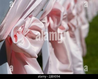 Reihe von weißen Stühlen geschmückt mit großen rosa Bändern bereit für eine Sommerhochzeit im Freien. Stockfoto