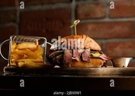 Pastrami-Sandwich auf Roggenbrot, serviert mit Gurken und Senfsoße. Leckeres Sandwich mit pommes Frites-Kartoffeln auf dunklem Hintergrund. Fastfood-Mittagessen Stockfoto