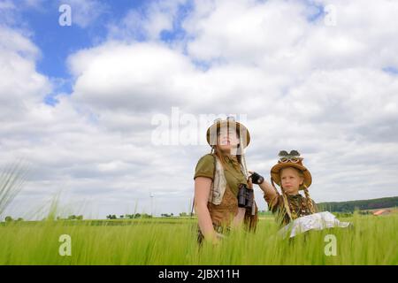 Zwei junge Mädchen werden in grünen Feldern gesehen, die die Landschaft beobachten. Mit Safarihüten und Khaki-Outfits bekleidet, tragen sie Ferngläser um den Hals Stockfoto