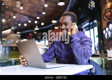 Positiver afroamerikanischer Telearbeiter, der auf einem Stuhl am Tisch sitzt und auf dem Smartphone spricht, während er im Café an einem Netbook fernarbeitet Stockfoto