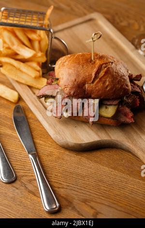 Pastrami-Sandwich auf Roggenbrot, serviert mit Gurken und Senfsoße. Leckeres Sandwich mit pommes Frites-Kartoffeln auf dunklem Hintergrund. Fastfood-Mittagessen Stockfoto