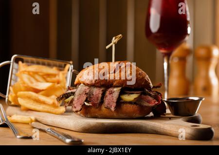 Pastrami-Sandwich auf Roggenbrot, serviert mit Gurken und Senfsoße. Leckeres Sandwich mit pommes Frites-Kartoffeln und Sangria-Getränk auf dunklem Hintergrund. Stockfoto