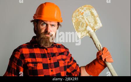 Seriöse Erbauer in Harthut mit Schaufel. Bärtiger Arbeiter in Arbeitshandschuhen und Helm mit Spaten. Stockfoto
