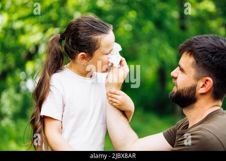 Seitenansicht des bärtig-hübschen Mannes im mittleren Alter, Vater, der mit einem weißen Taschentuch die Nase seiner Töchter abwischt und im Wald spaziert. Stockfoto