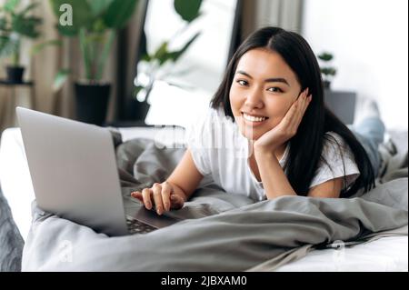 Hübsches, positives asiatisches Brünette-Mädchen liegt zu Hause auf dem Bett, nutzt Laptop, durchsucht soziale Netzwerke, chattet online mit Freunden, schaut sich einen Film an, entspannt sich am Wochenende zu Hause, schaut in die Kamera, lächelt Stockfoto