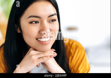 Nahaufnahme eines schönen positiven Brünette moderne braune Augen Mädchen asiatischer Nationalität, in lässiger Kleidung, Blick zur Seite, drinnen, freundlich lächelnd, träumen, denken Stockfoto