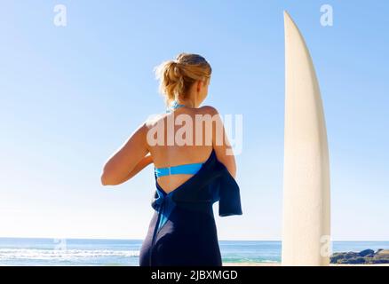 Rückansicht einer jungen Frau, die neben ihrem Surfbrett steht und ihren Neoprenanzug anlegt Stockfoto