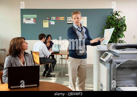 Mann an der Kopiermaschine im Gespräch mit einer Kollegin auf dem Laptop Stockfoto