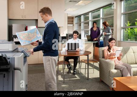 Eine Gruppe von Personen, die in und im Büro an Tablets und Laptops arbeiten, ein Mann, der Kopien am Drucker anfertigte. Stockfoto