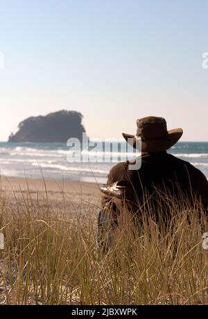 Rückansicht eines Mannes, der einen akubra-Hut und Schuhe trägt, die über seine Schulter hinausgezogen sind und am Strand sitzt und auf das Meer blickt Stockfoto