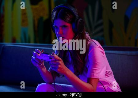 Frau mit Gamecontroller sitzt vor dem Fernseher und trägt ein Mikrofon-Headset mit Intensität. Stockfoto