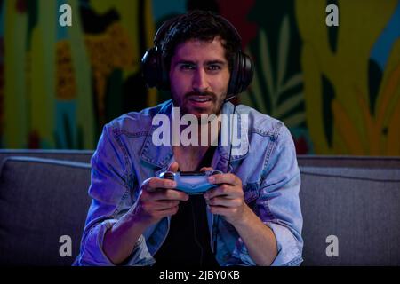 Junger Mann spielt nachts in seinem Wohnzimmer Videospiele mit Gamecontroller und Kopfhörern. Stockfoto