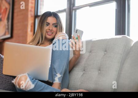 Glückliche hispanische Frau, die zu Hause auf dem Mobiltelefon mit einem Laptop spricht Stockfoto