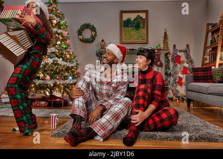 Vorderansicht eines gemischten Rennpaares mit ihrer kleinen Tochter zu Weihnachten, das Weihnachtsmützen trägt, die zusammen auf dem Boden sitzen und Geschenke öffnen. Stockfoto