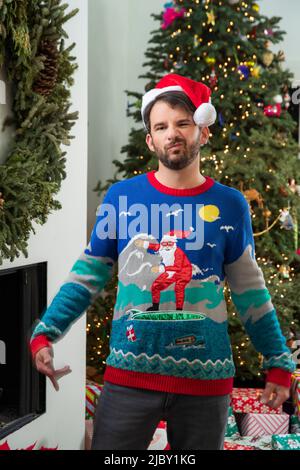 Junger Mann, der vor dem Weihnachtsbaum steht, trägt einen hässlichen Weihnachtspullover und einen roten Weihnachtsmütze. Stockfoto
