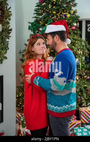 Liebevolles Paar in hässlichen Weihnachtspullis, die sich neben dem Baum umarmen. Stockfoto