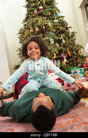 Porträt eines Mädchens, das vor dem Weihnachtsbaum sitzt und ihren Vater spielt. Stockfoto