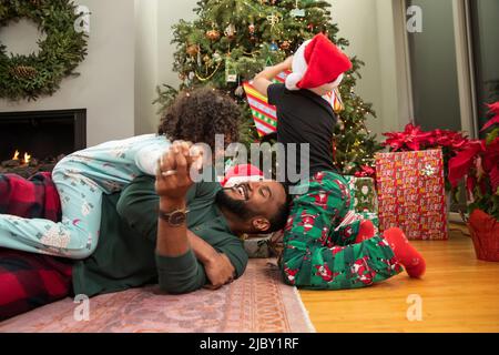 Fröhlicher Vater kämpft mit seiner Tochter am Boden, während sein Sohn Weihnachtsgeschenke öffnet. Stockfoto