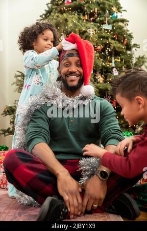 Mann, der vor dem Weihnachtsbaum sitzt und von seinen Kindern geschmückt wird Stockfoto