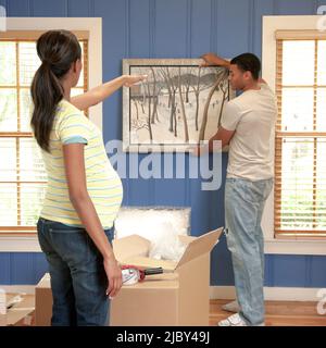 Ein afroamerikanisches Paar zieht zusammen ins Haus, während die Schwangere zeigt, wo sie die Malerei aufhängen kann, Stockfoto