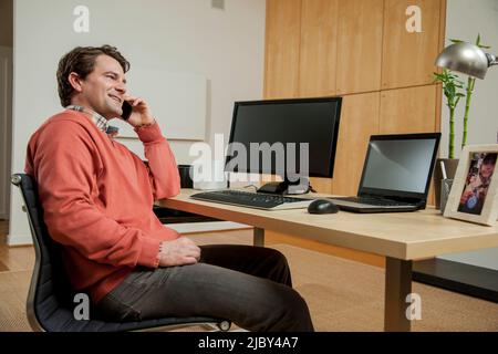 Mann, der im Heimbüro sitzt und sich auf dem Stuhl zurücklehnt und am Handy redet. Stockfoto