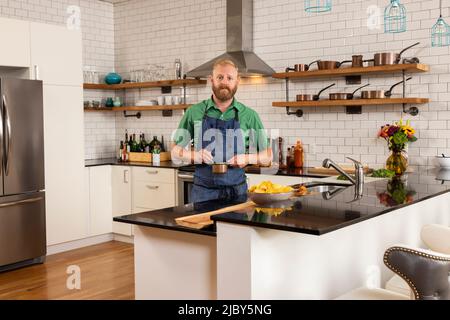 Porträt eines kaukasischen Mannes mit Bart, der in der heimischen Küche mit Schürze steht und in die Kamera schaut. Stockfoto