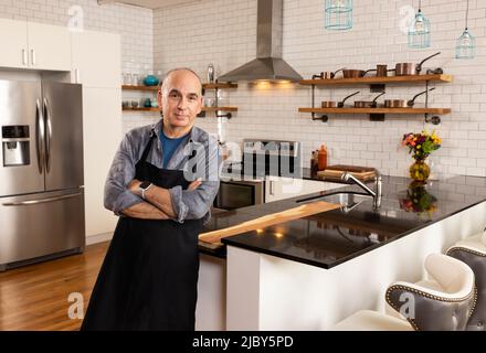 Mann lehnte sich mit gekreuzten Armen an der Küchenarbeitspe und schaute in die Kamera. Stockfoto