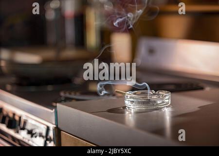 Startseite Interieur, Detail von Aschenbecher, der eine beleuchtete CBD-Pre-Roll-Joint hat, Zigarette mit Rauch steigt in der Küche neben Herd sitzen Stockfoto