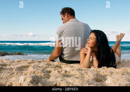 Mann und Frau entspannen sich am Strand, Mann spielen mit Sand und Frau liegen auf dem Bauch mit gekreuzten Beinen hinter ihr Stockfoto
