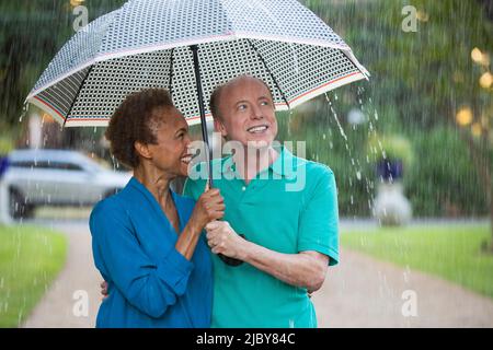 Ein älteres Paar, das im Regen durch den Park mit einem Regenschirm ging, und ein Mann, der auf das Wetter schaute Stockfoto