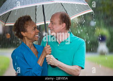 Älteres Paar, das im Regen mit Sonnenschirm durch den Park läuft und sich liebevoll anschaut Stockfoto