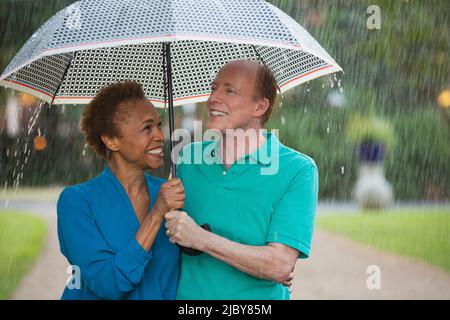 Älteres Paar, das im Regen mit Sonnenschirm durch den Park läuft und sich liebevoll anschaut Stockfoto