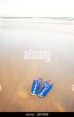 Jandals, Tangas, mit australischer Flagge auf dem Sand und Wellen im Hintergrund Stockfoto