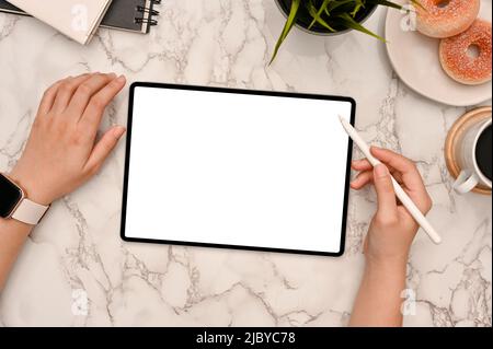 Über der Ansicht zeichnet und skizziert Eine Frau ihr Design auf einem Tablet mit einem Eingabestift auf ihrer modernen, stilvollen Marmorplatte. Modell mit weißem Bildschirm des Tablets. Stockfoto