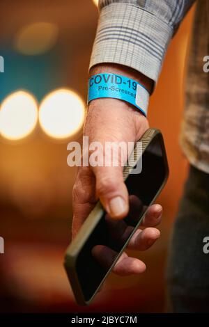 Nahaufnahme einer Männerhand, die ein COVID-19 vorabgeschirmtes Armband trägt und ein Mobiltelefon hält. Stockfoto
